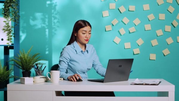 Asian administrative assistant works on a laptop at her desk. Office setting with sticky notes on the wall.