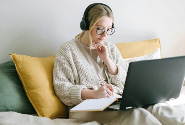 Woman with glasses working on laptop in bed. Headphones, notebook, and pen. Remote work lifestyle.
