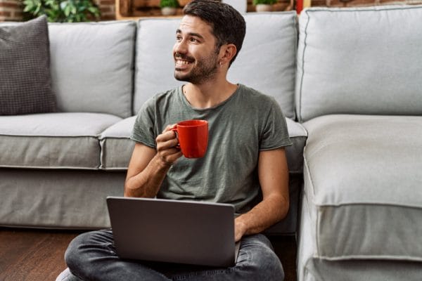 Adult learning mathematics online. Man with laptop and coffee cup smiles, seated on floor.