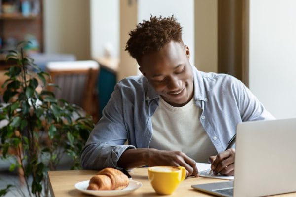 Call centre administrator working. Man writing at desk with laptop, coffee, and croissant. Office admin.