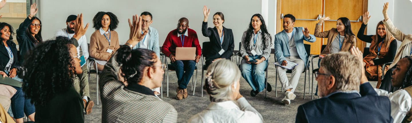 Diverse group of students and facilitators participating in a college seminar or workshop, engaging actively with raised hands and lively discussion, promoting learning and collaboration.